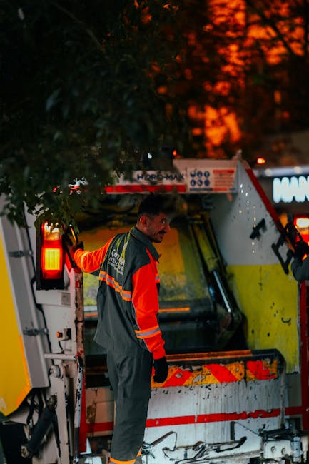 A waste collection worker dressed in green uniform and safety helmet stands on the rear step of a compact rubbish collection van, which is parked on a residential street lined with trees and parked ca