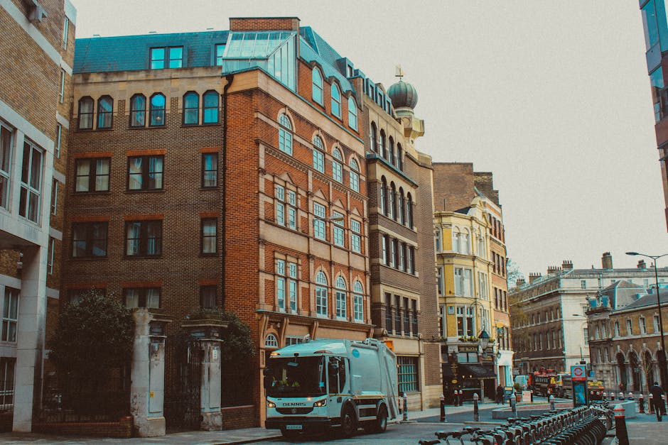 A large yellow waste collection truck with a hydraulic lifting mechanism is parked on a city street adjacent to a modern multi-storey building with large glass windows and horizontal metal paneling. T