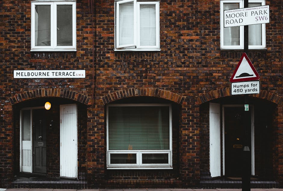 A brick residential building with a dark red and brown textured facade featuring three white-framed windows on the upper floor, one of which is open with partially visible blinds inside, and three arc