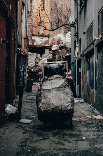 A narrow urban alleyway with uneven, dark asphalt ground cluttered with scattered debris and small pieces of waste. In the center, a large, black wheeled sack made of durable material is positioned up